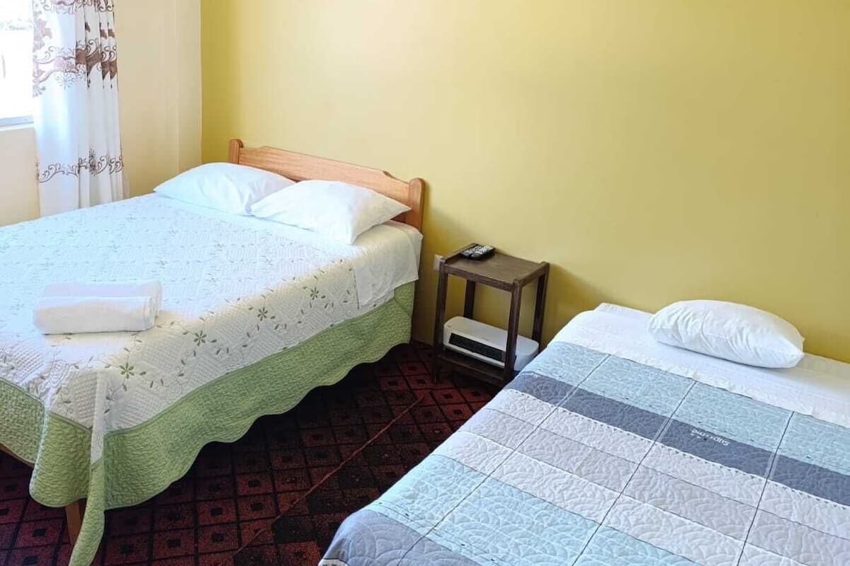 A room with two beds, one with a green bedspread and the other with a blue and white quilt. There is a small wooden nightstand with a remote control on it.