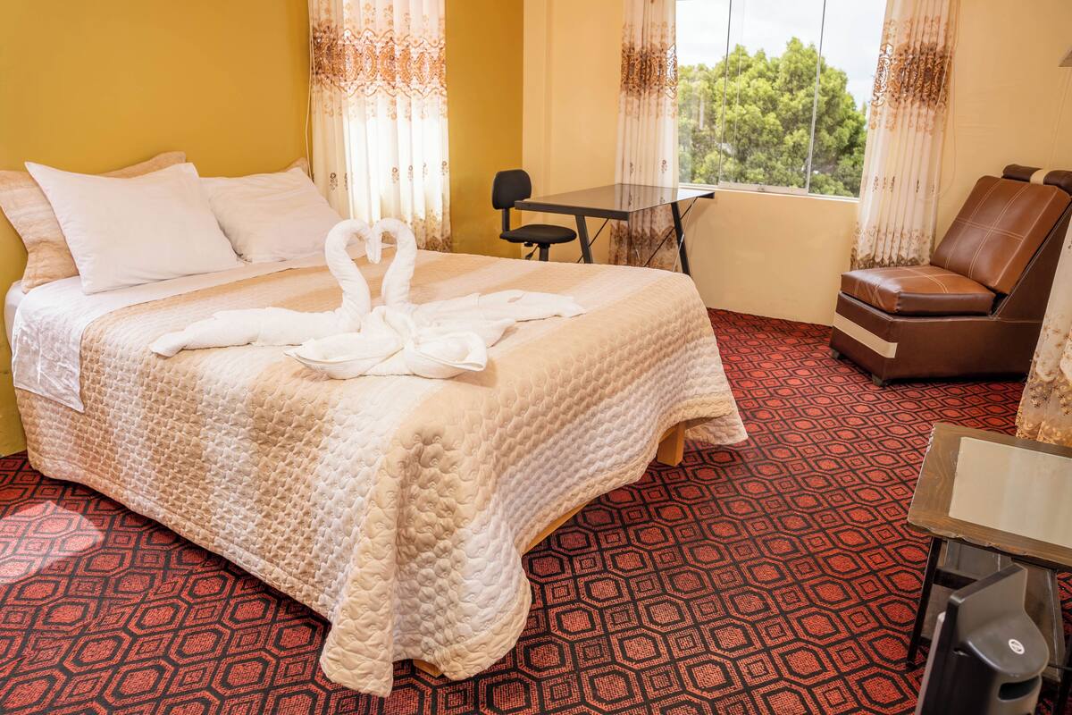 A hotel room with a bed, a chair, a table, and a view of trees through the window.