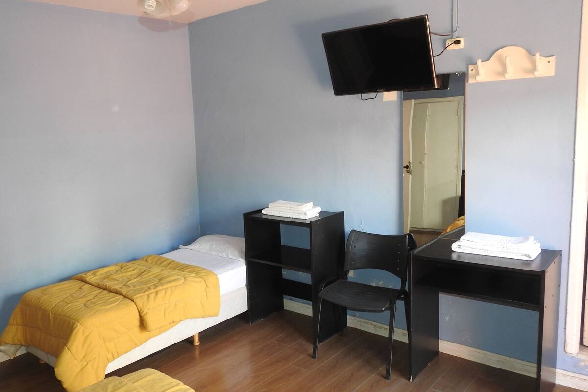 Quarto pequeno com uma cama de solteiro, piso de madeira, escrivaninha, cadeira e televisão fixada na parede.
