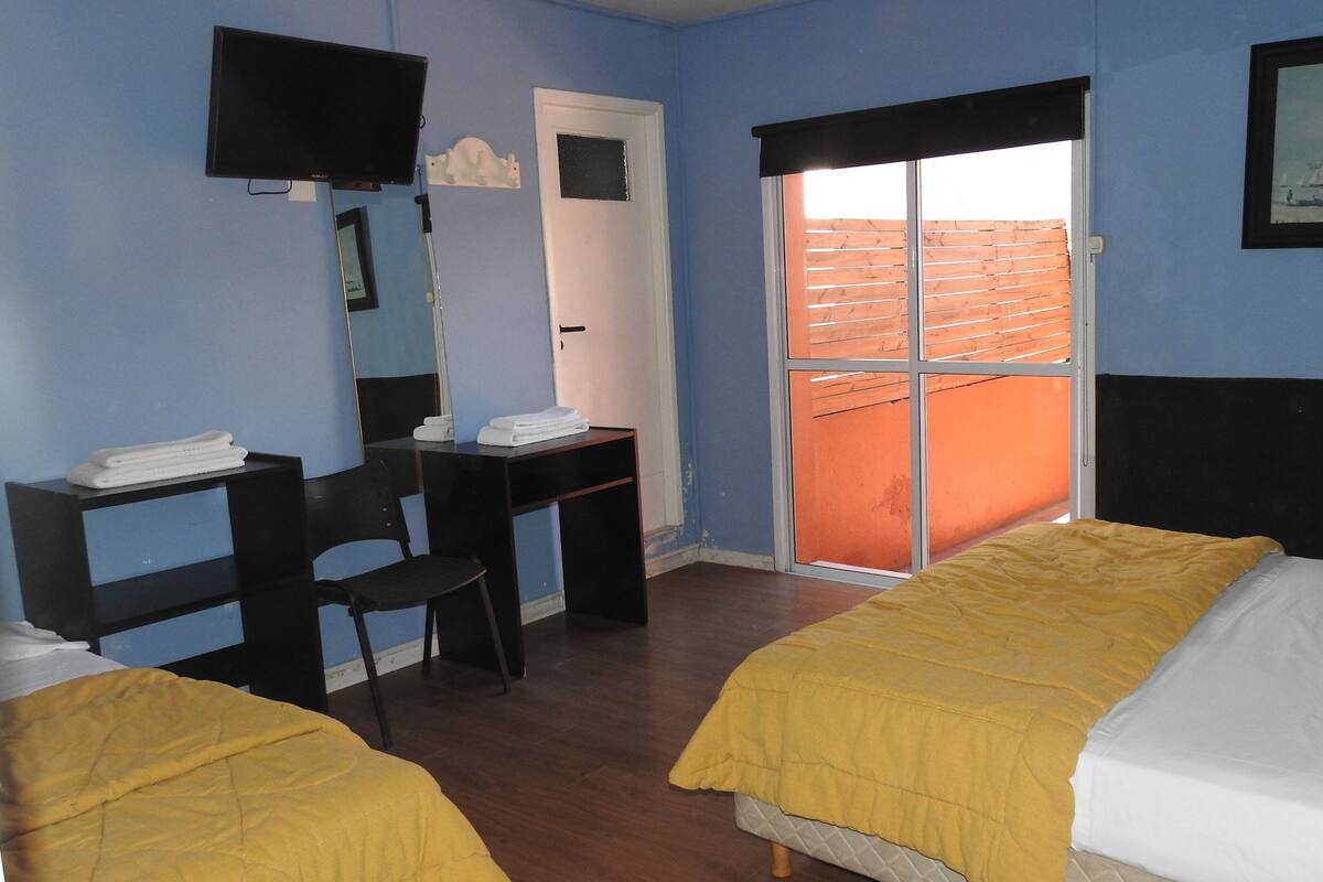 Quarto de hotel com duas camas, uma escrivaninha, uma cadeira, uma televisão e uma janela com vista para um edifício vermelho.