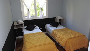 Ein kleines Zimmer mit zwei Betten, jeweils mit einer gelben Decke, einem Nachttisch mit Telefon und einem Fenster mit weißen Vorhängen.