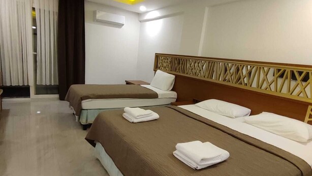Quarto de hotel com três camas, cada uma com colcha marrom e almofadas brancas, e cabeceira com padrão geométrico.