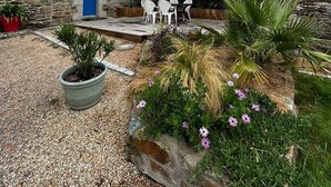 Un bâtiment en pierre avec une porte bleue, une terrasse dotée de mobilier d’extérieur et un jardin où poussent différentes plantes et fleurs.