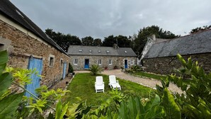 Un bâtiment en pierre avec une cour verdoyante, deux portes bleues et des fauteuils de jardin blancs.