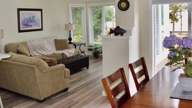 A living room with a sofa, a dining table, and chairs. There is a window with blinds and a clock on the wall.