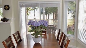 A dining room with a wooden table and chairs, a potted plant, and a view of the outdoors through large windows.