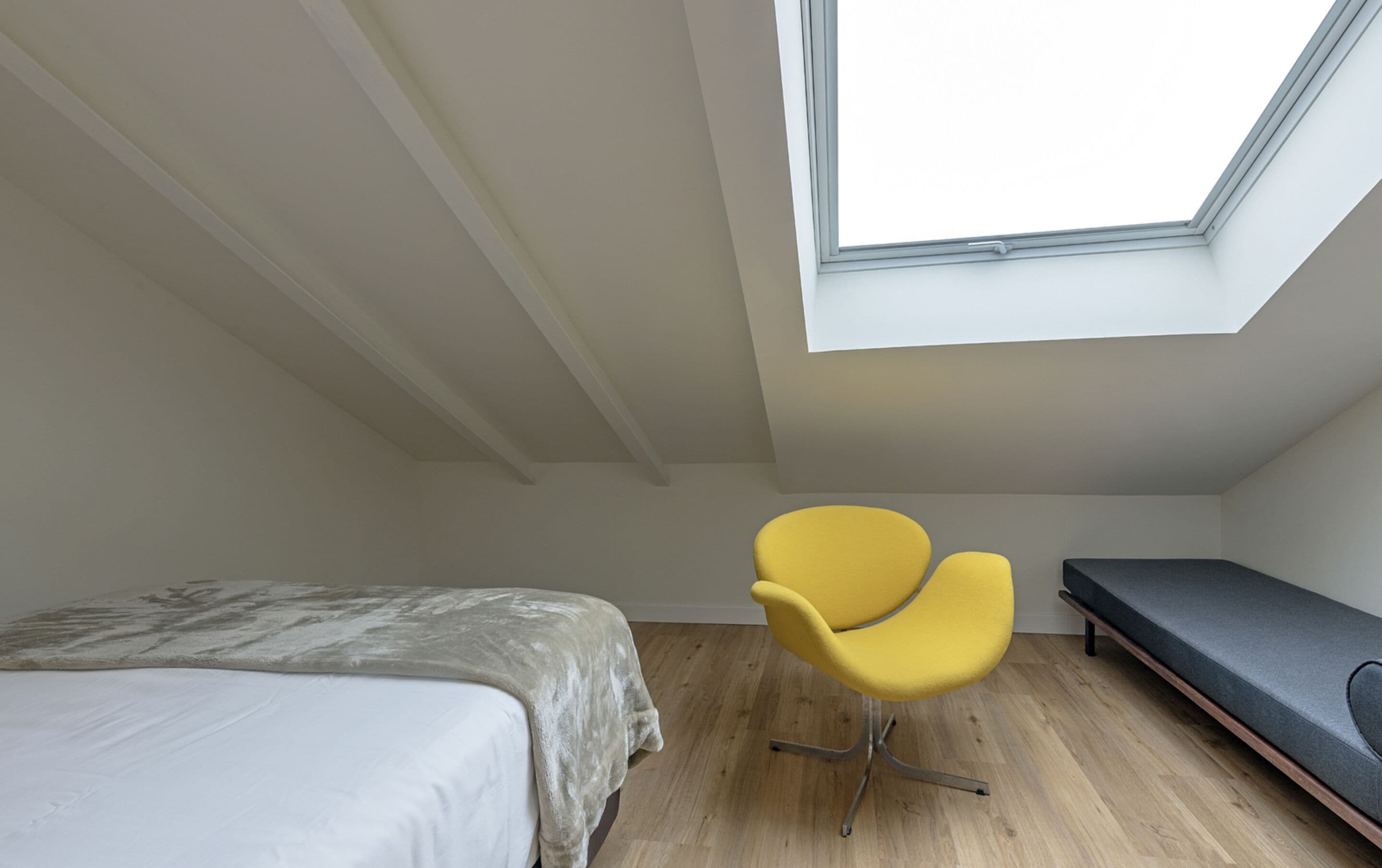 A small, modern room with a skylight, a bed, a yellow chair, and a bench.