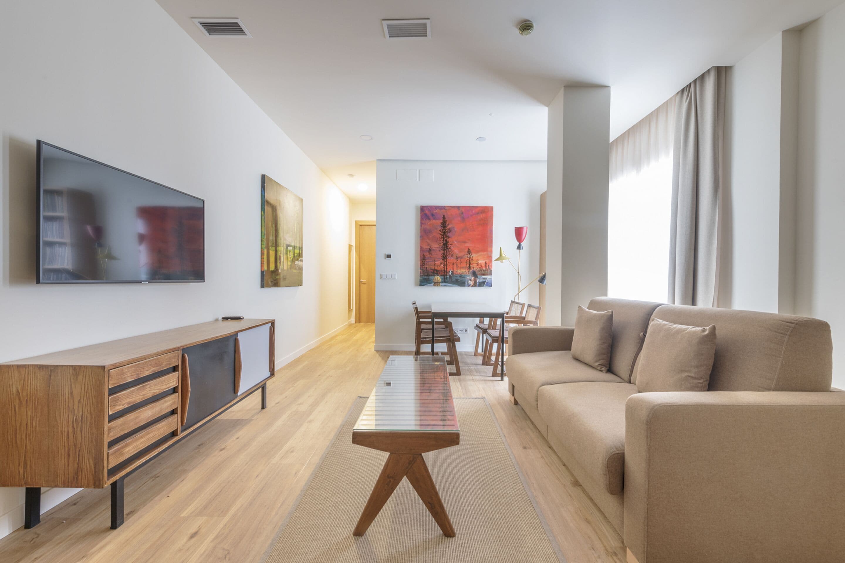 Una sala de estar moderna con un sofá, una mesa de centro y un televisor montado en la pared. En el fondo, se observa un comedor con una mesa y sillas.