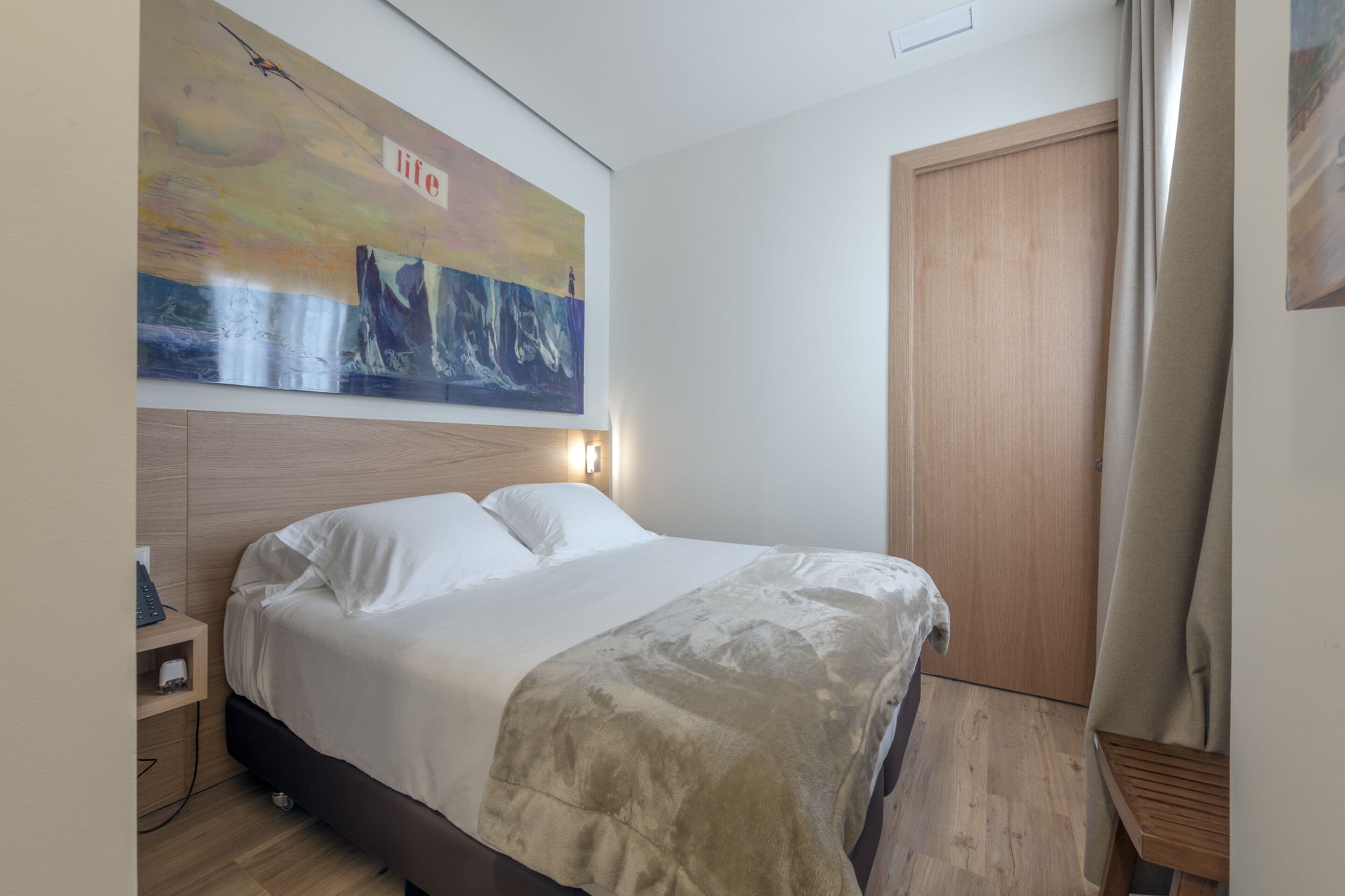 Una cama bien hecha con cabecera, una mesita de noche con lámpara y un cuadro abstracto grande en la pared.