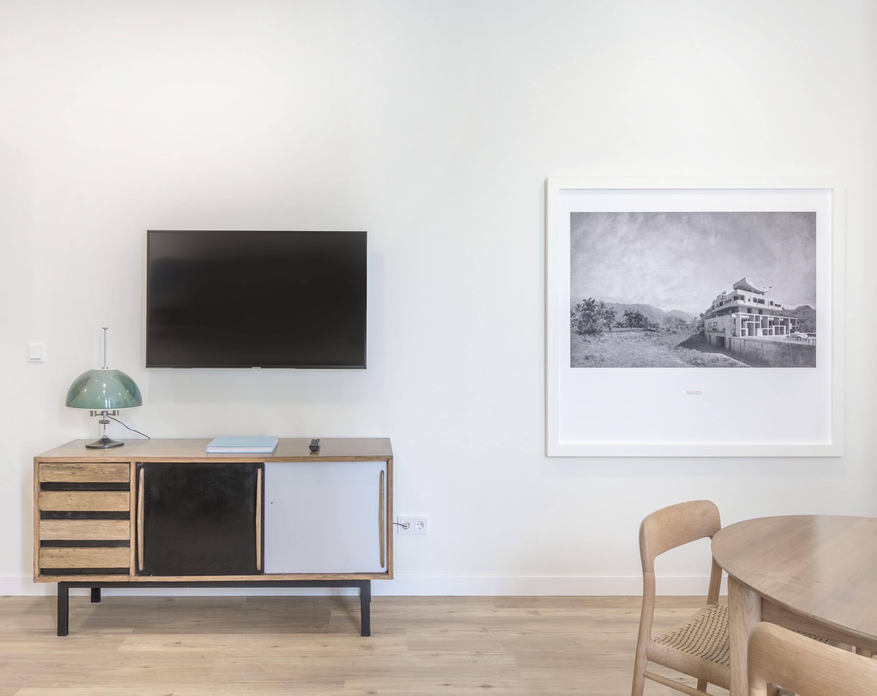 Una sala de estar minimalista con un televisor de pantalla plana montado en la pared, un mueble de madera con cajones, una lámpara de escritorio verde y una fotografía en blanco y negro enmarcada en la pared.