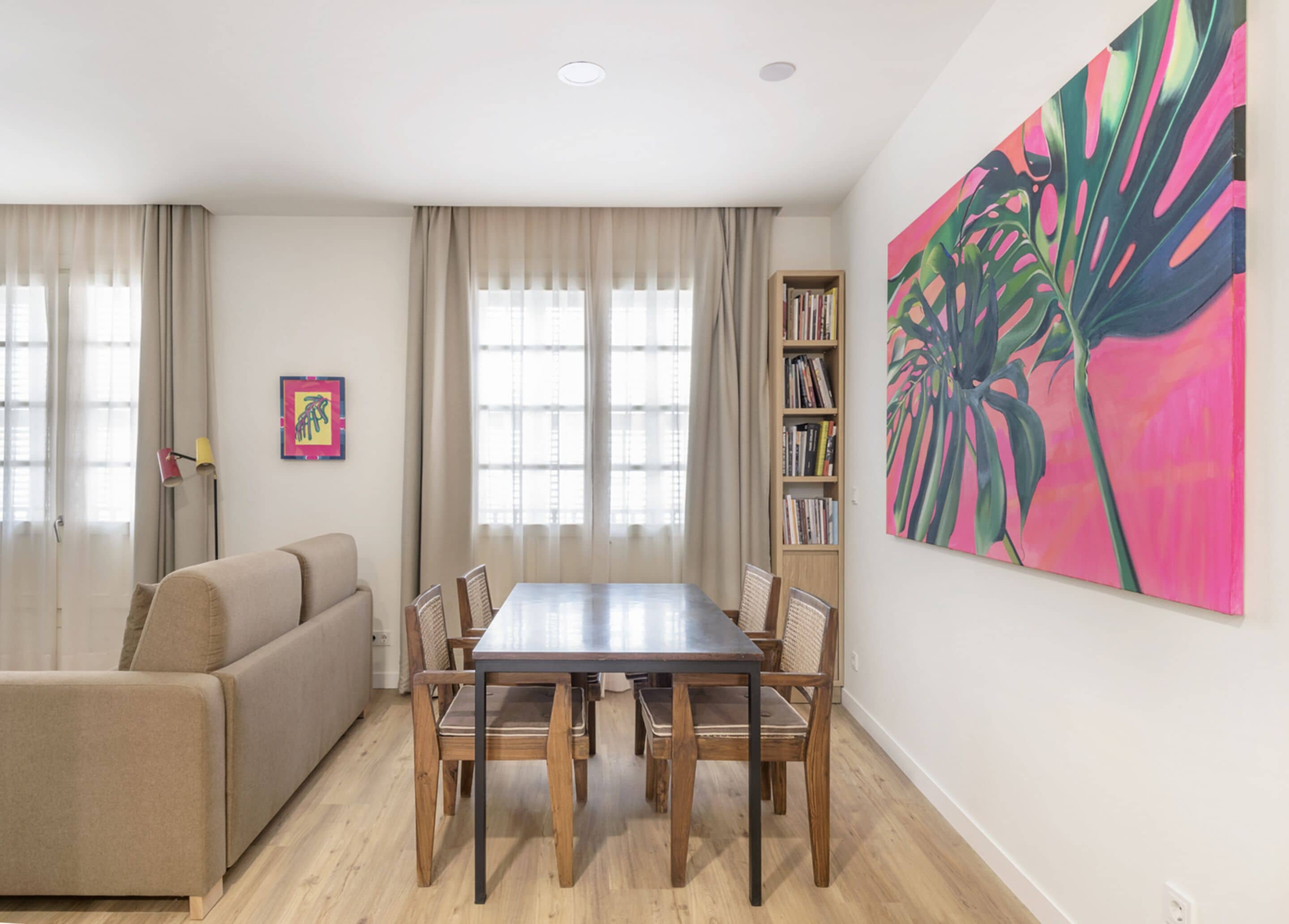 Una sala de estar con un sofá color crema, una mesa de comedor de madera con sillas, una estantería y un cuadro grande en la pared.