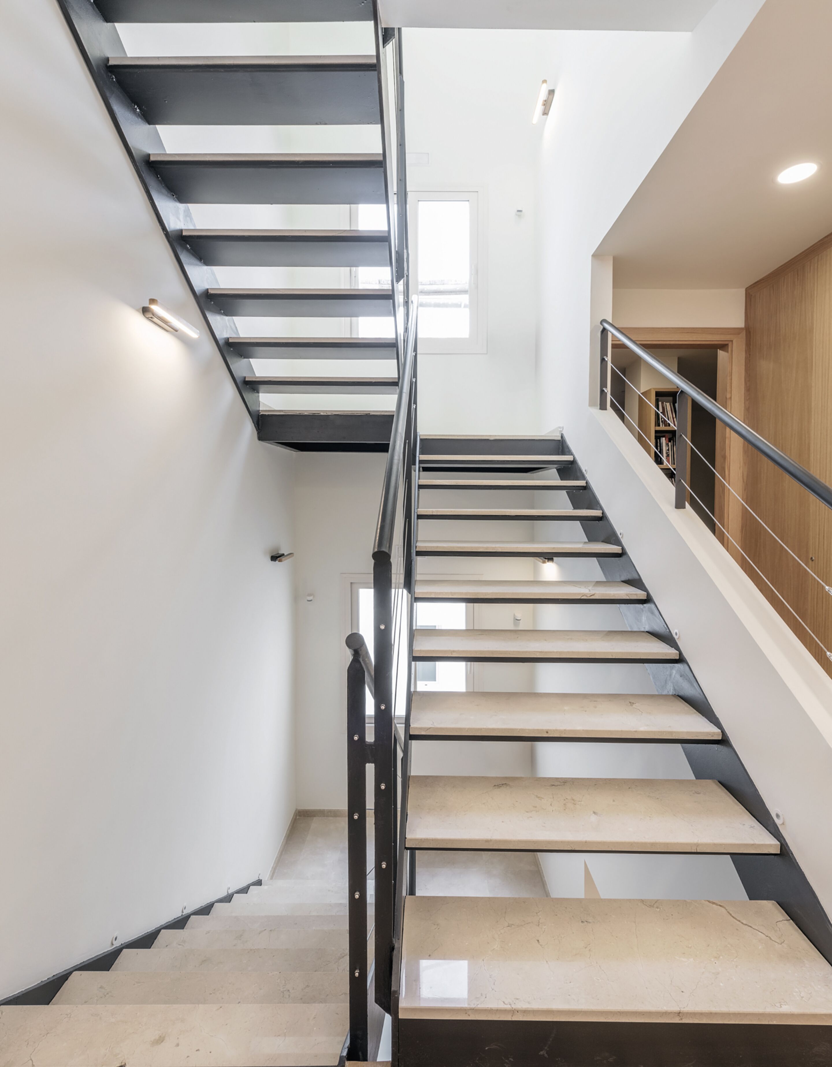 Una escalera moderna con barandillas de metal y peldaños de madera, que conduce a un rellano con una puerta de madera y una estantería.