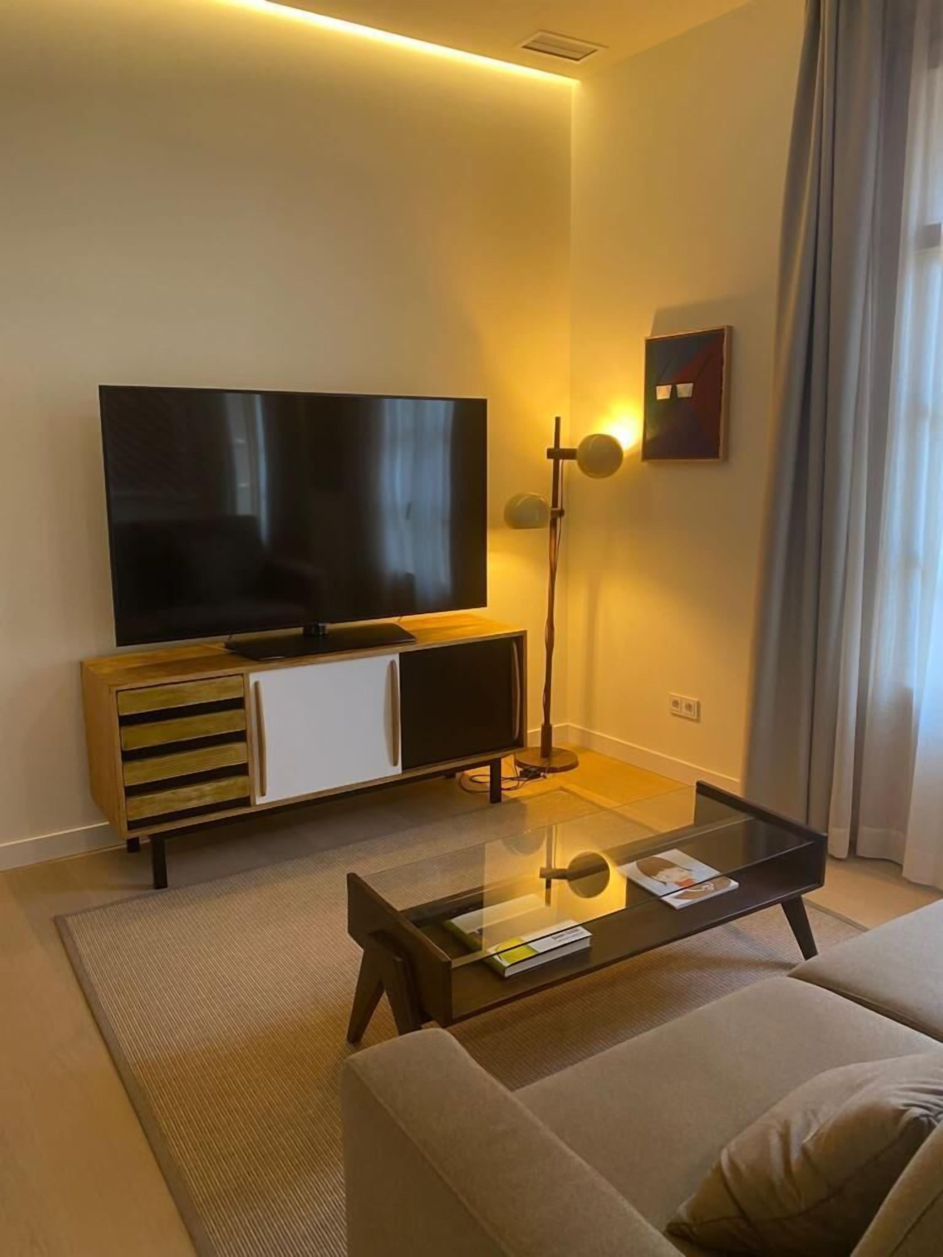 Un saló modern amb televisió de pantalla plana, un moble de fusta, una làmpada de peu i una taula de centre de vidre.