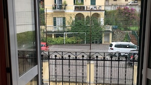 A view through a window of a building with a sign reading "OPERE PARROCCHIALI" and a fenced courtyard with a parked car.