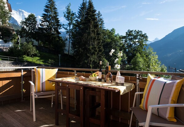 Comedor al aire libre con mobiliario de madera y una mesa preparada para dos, con vistas a un paisaje montañoso.