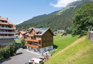 A modern two-story building with a wooden facade, a balcony with outdoor seating, and a parking area in a mountainous area. - Le Manoir Penthouse - spacious, new and central location (Wengen)