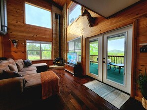 Een knus blokhutinterieur met een open haard, houten wanden, een bank en een schuifdeur naar een terras.