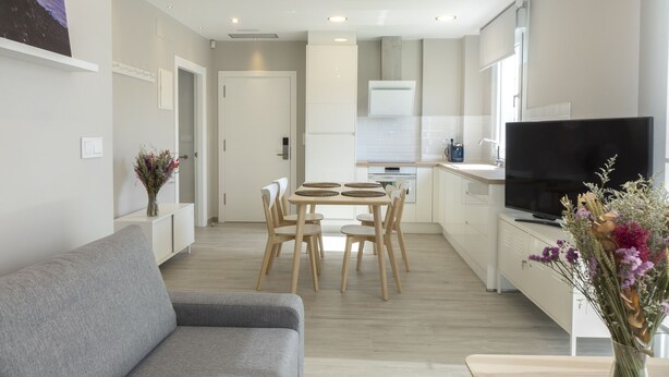 A modern living room with a grey sofa, a dining area with a wooden table and chairs, a kitchen with white cabinets and a flat-screen TV. - Two bedroom apartment with garden - DDS ONS (Vigo)