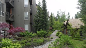 A modern building with a grey facade, a wooden deck, and a garden with various plants and trees.