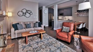 A modern living room with a sofa, coffee table, and a leather armchair. A kitchen is visible in the background with a dining area.