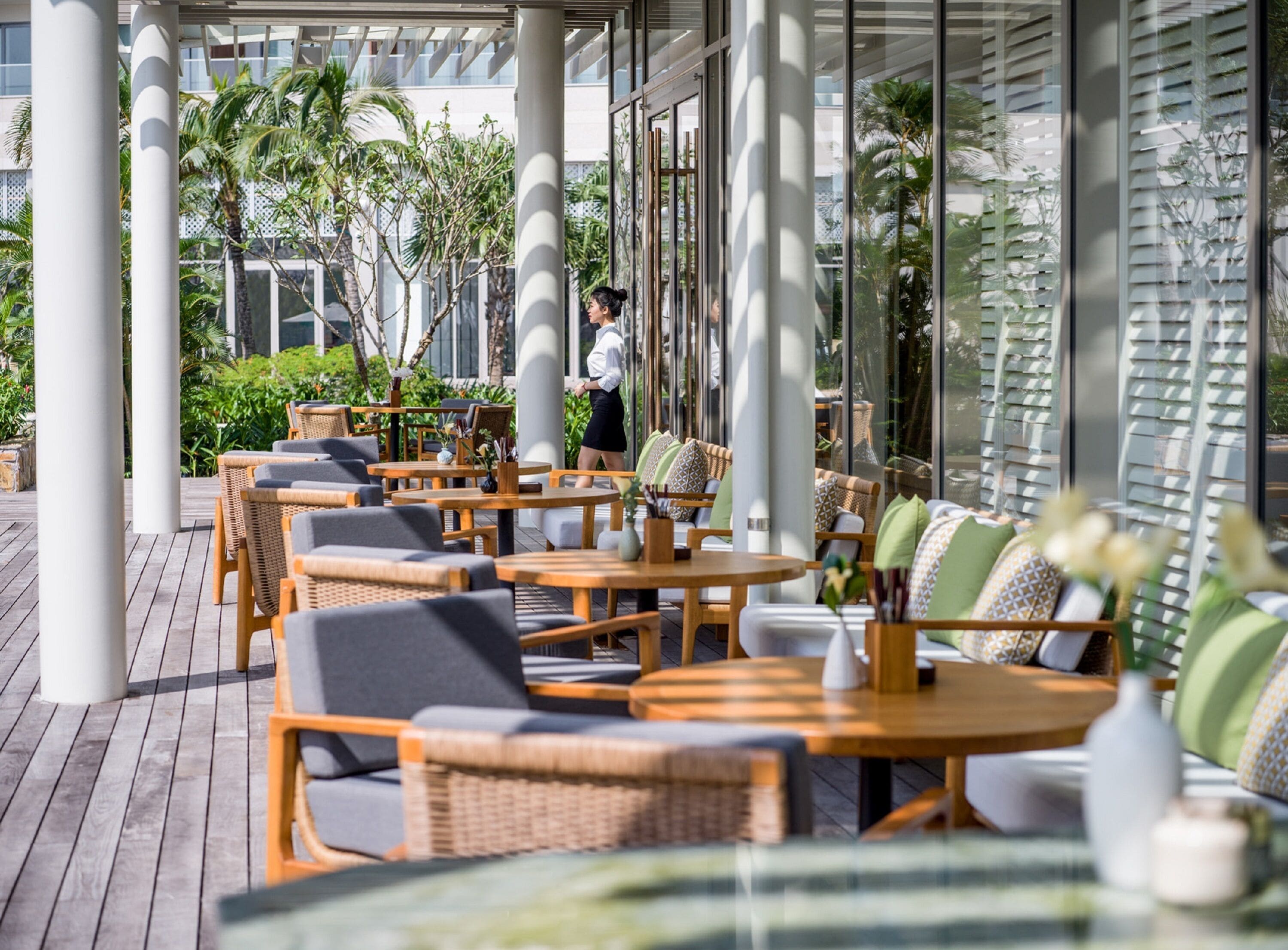 An outdoor seating area with wicker furniture and tables, surrounded by large windows and columns.