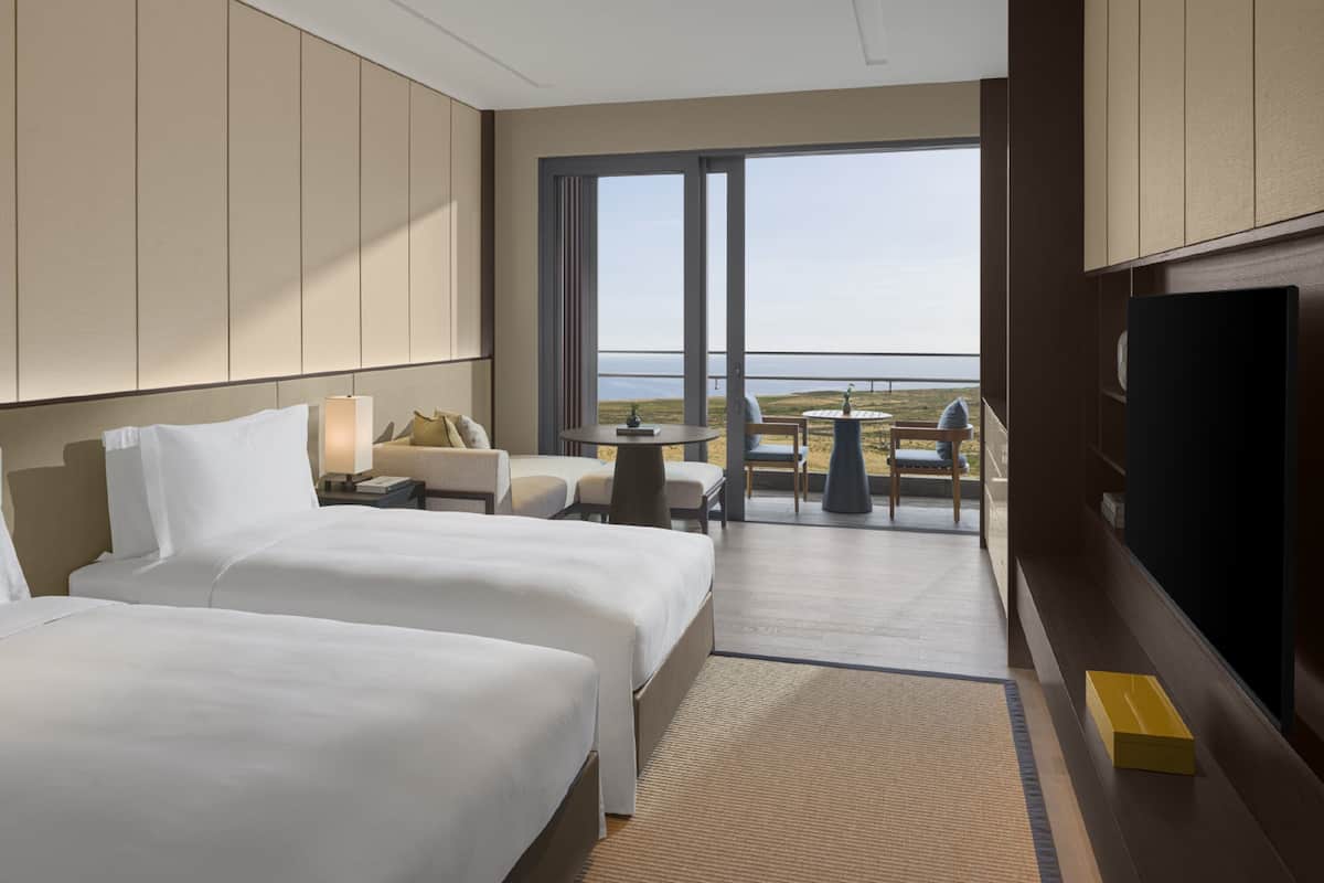 A hotel room with two beds, a TV, a balcony with a table and chairs, and a view of the sea.