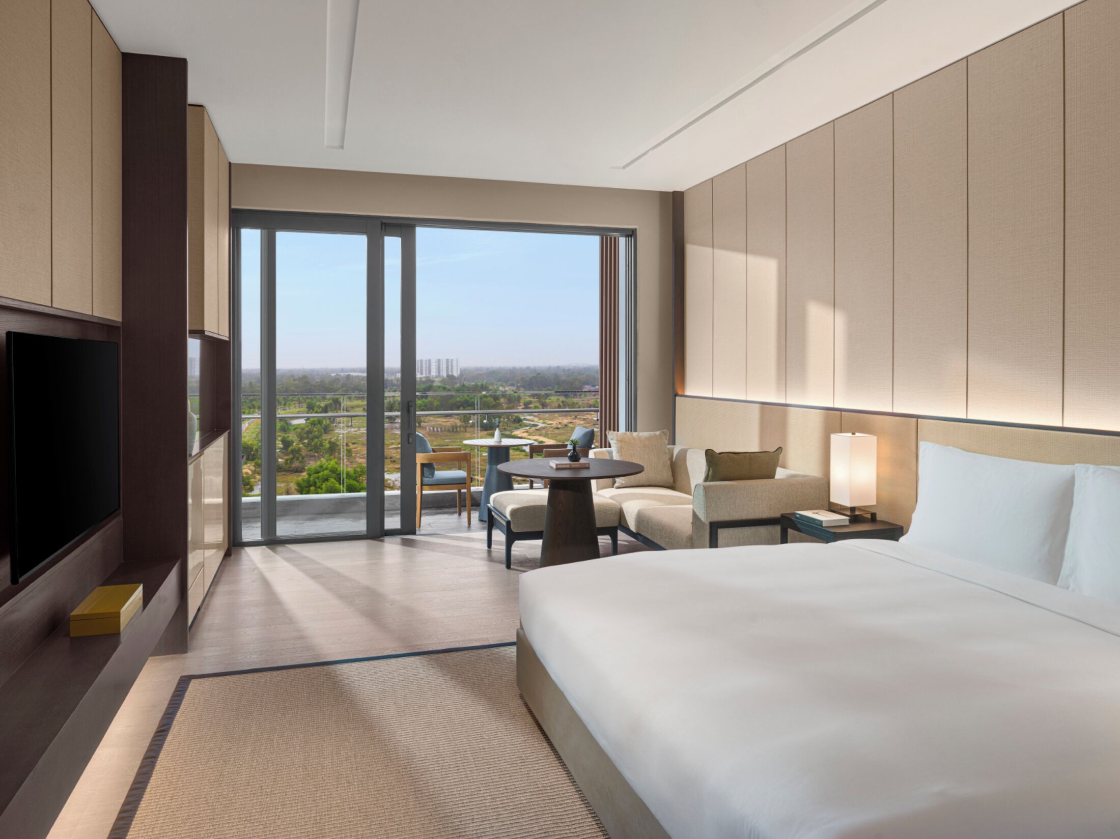 A hotel room with a large bed, a TV, a desk with a chair, a table with chairs, and a view of the cityscape.