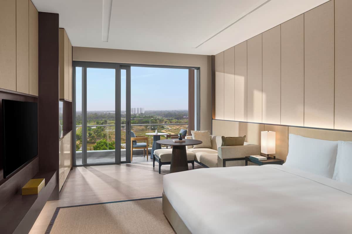 A hotel room with a large bed, a TV, a desk with a chair, a table with chairs, and a view of the cityscape.