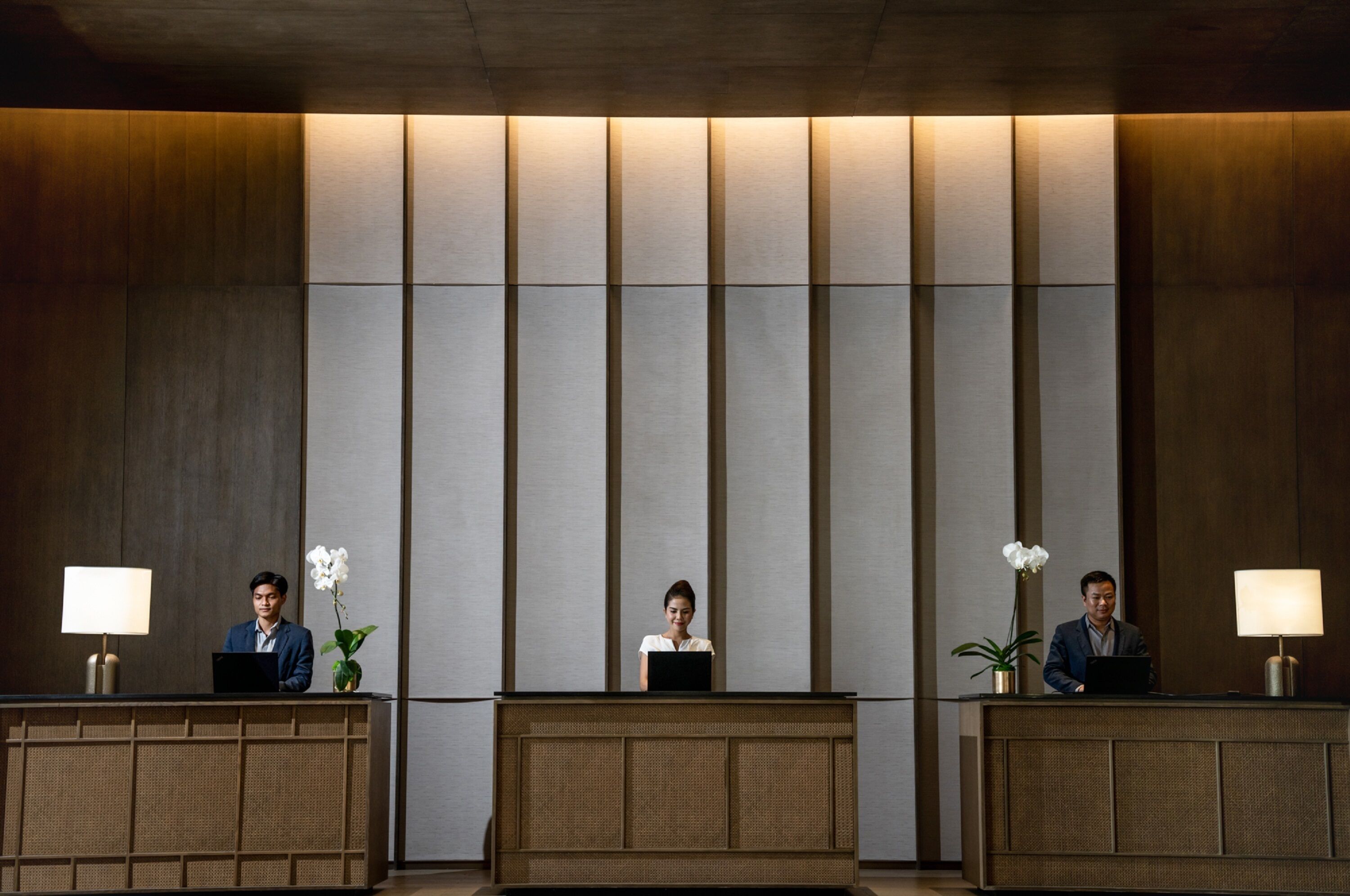 Una recepción con tres empleados, cada uno con un portátil, en un vestíbulo moderno con paneles de madera verticales y una paleta de colores neutros.