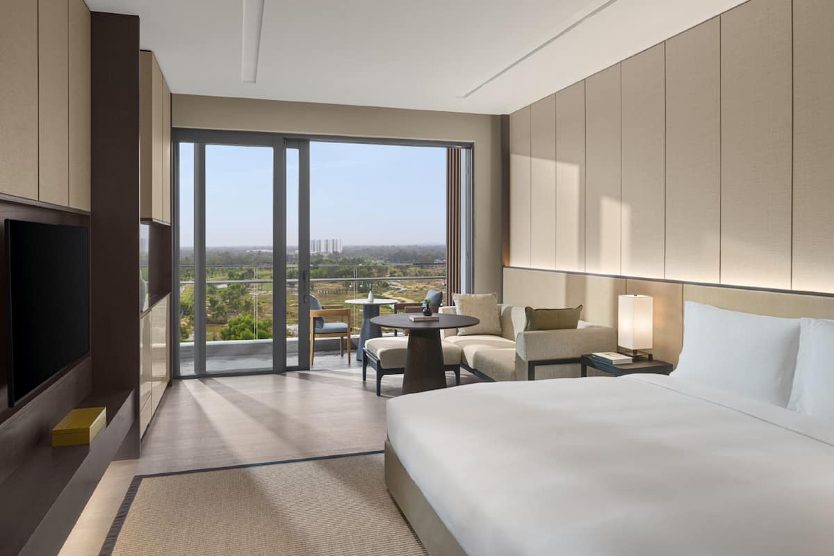 A modern hotel room with a large bed, a flat-screen TV, a dining area with a table and chairs, and a balcony with a view of the cityscape.