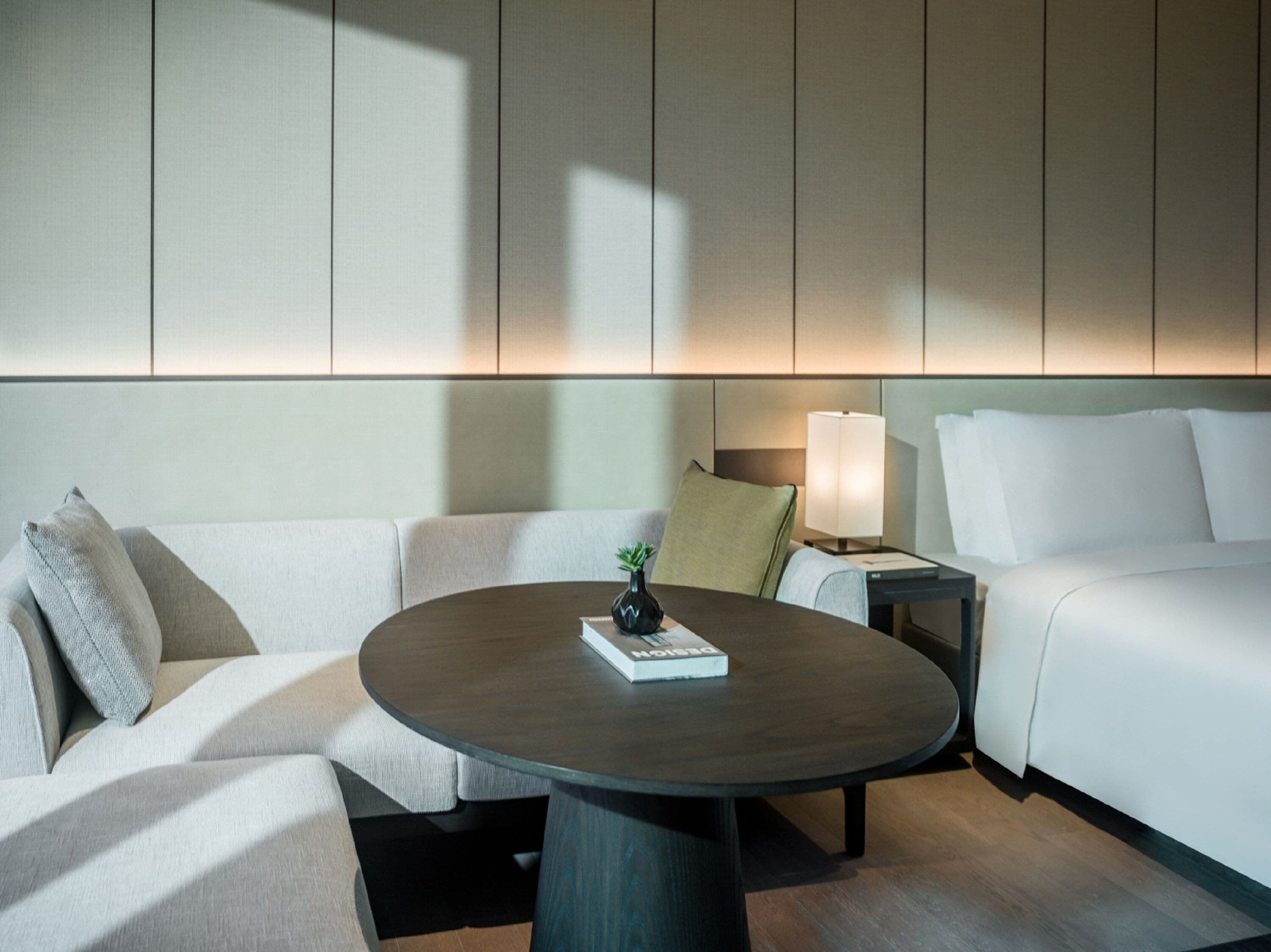 A modern hotel room with a round table, a sofa, a bed, and a nightstand with a lamp.
