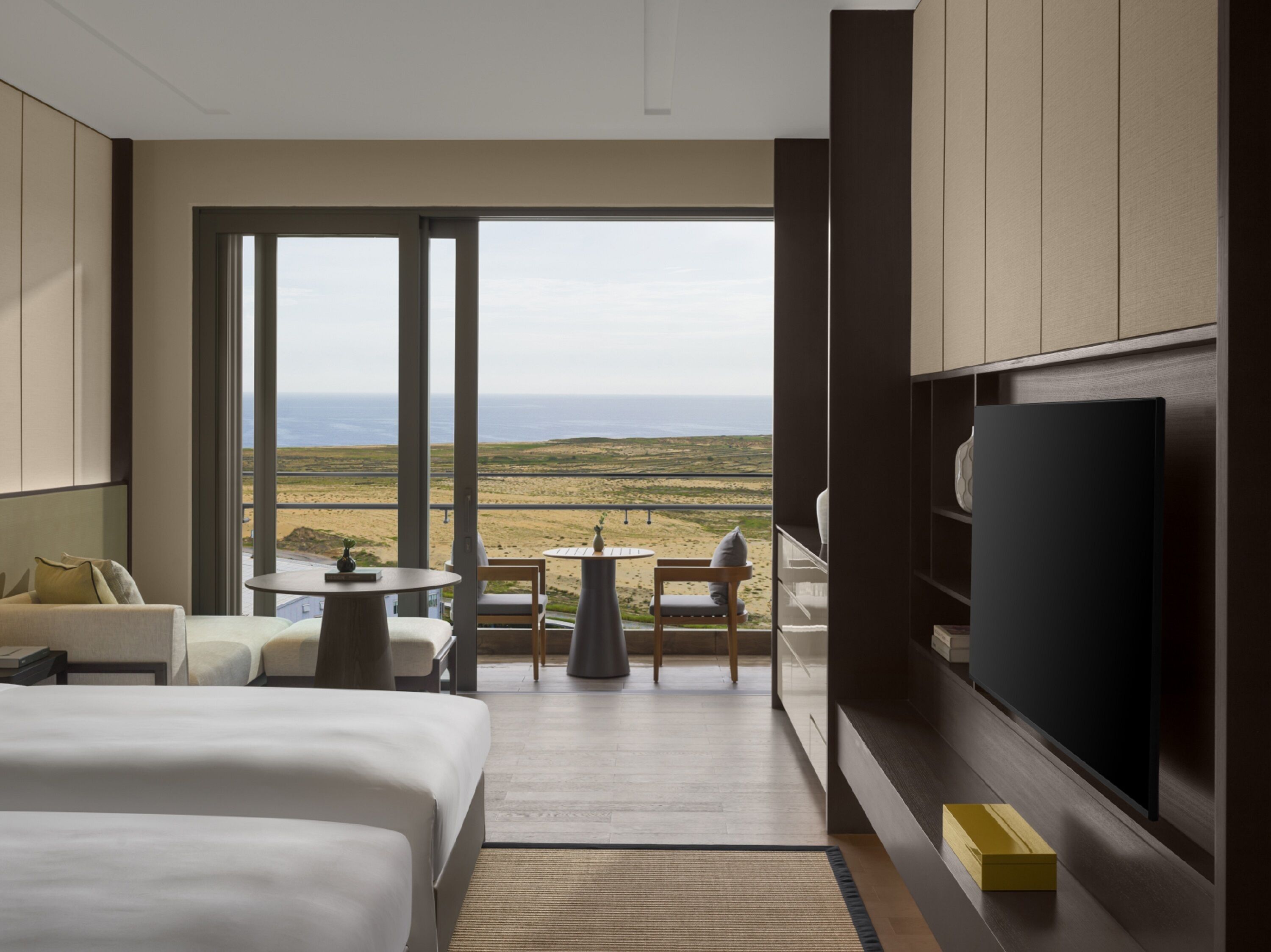 Ein modernes Hotelzimmer mit einem großen Bett, einem Fernseher, einem Balkon mit Blick auf den Strand und einem kleinen Tisch mit Stühlen.