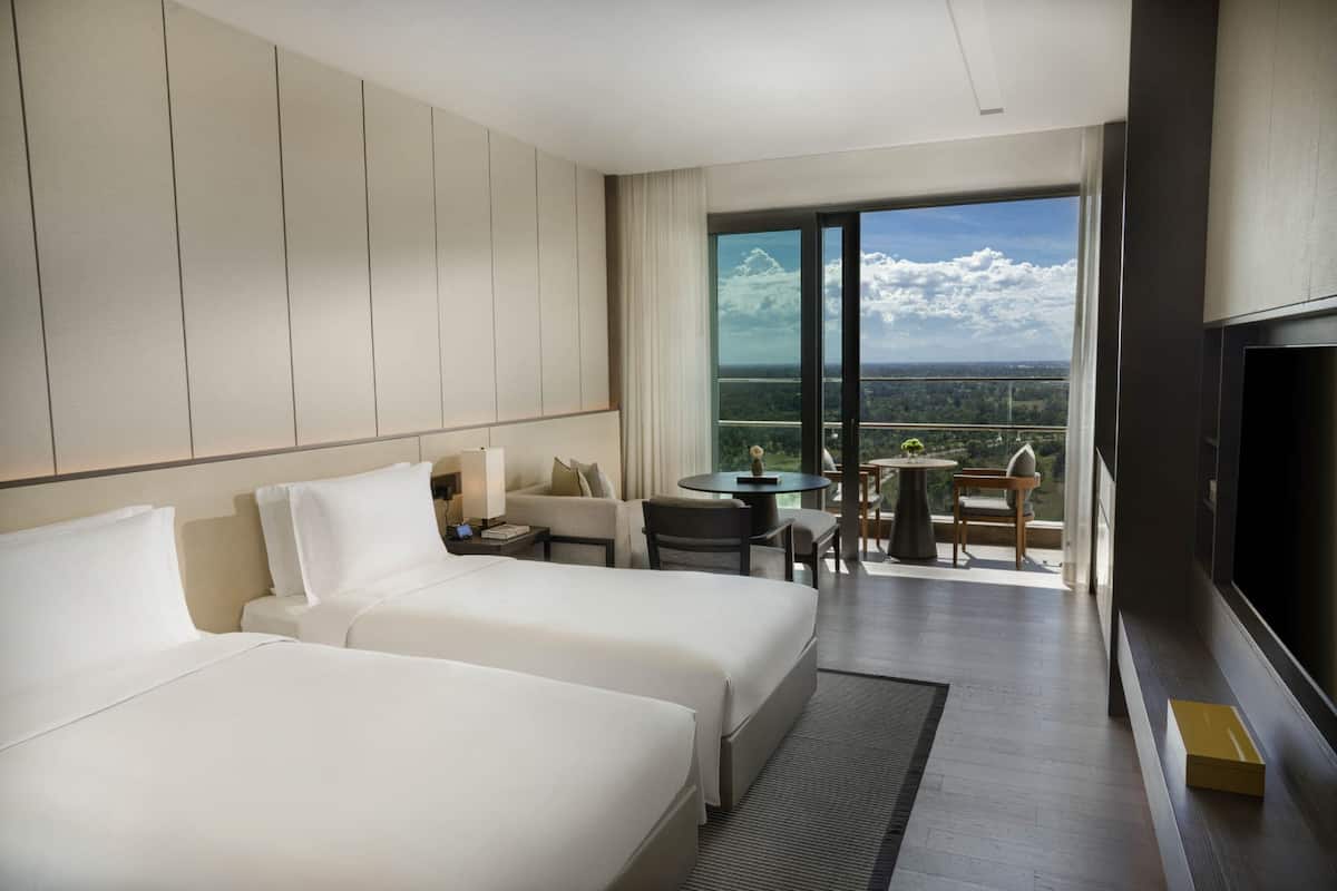 A hotel room with two beds, a TV, a balcony with a table and chairs, and a view of the city.