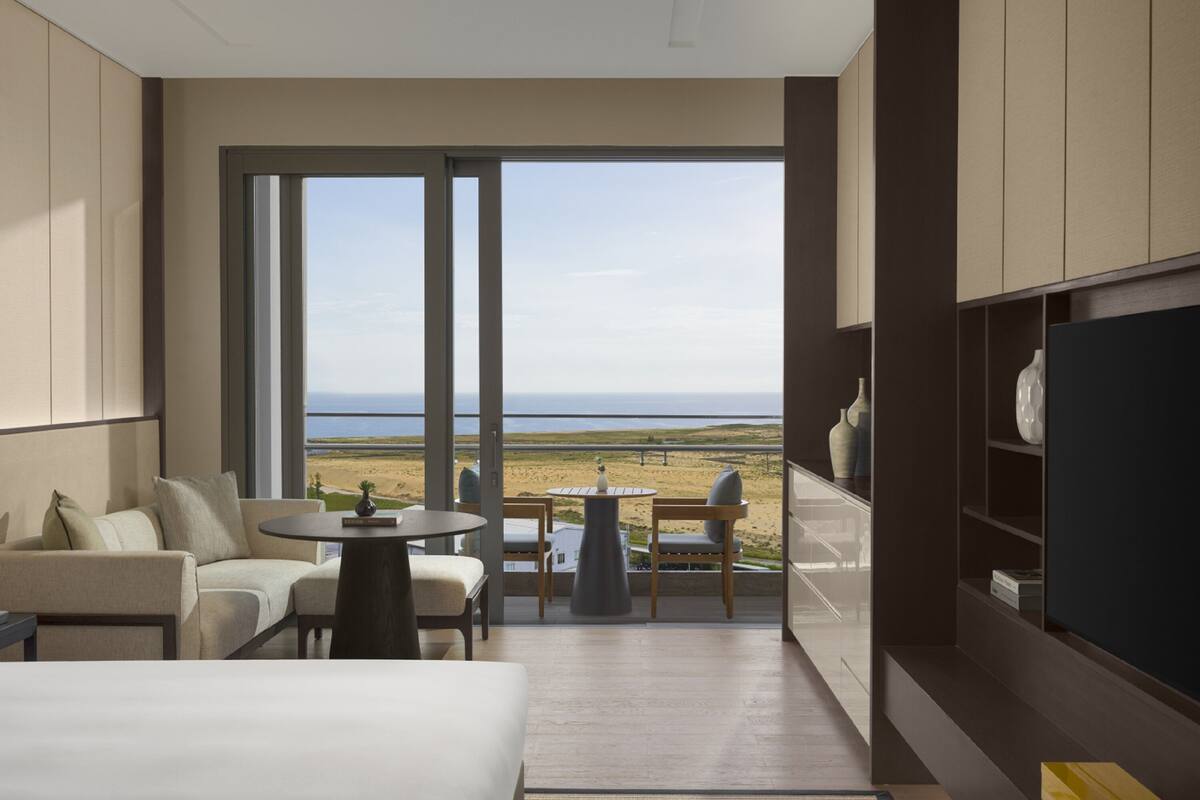 A modern hotel room with a large bed, a seating area with a table, and a view of the beach through large windows.