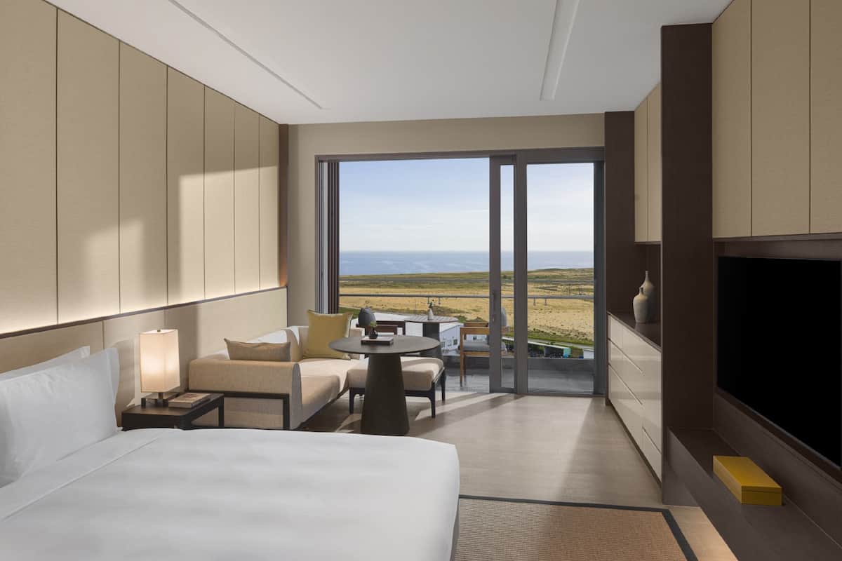 A modern hotel room with a large bed, a TV, a seating area with a table and chairs, and a balcony with a view of the beach.