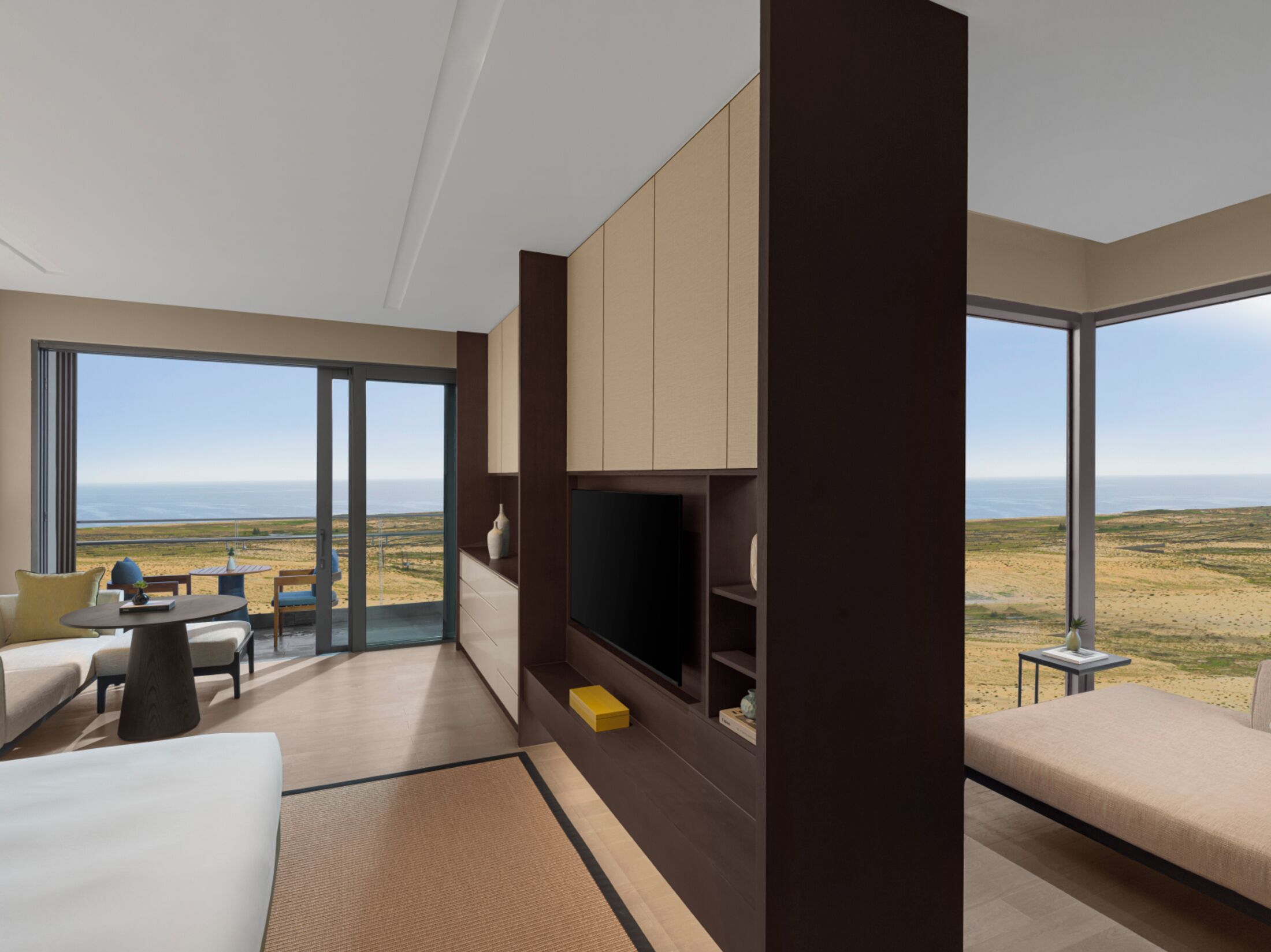 A modern hotel room with a large TV, comfortable seating, a sofa, a dining area, and a balcony with a view of the beach.