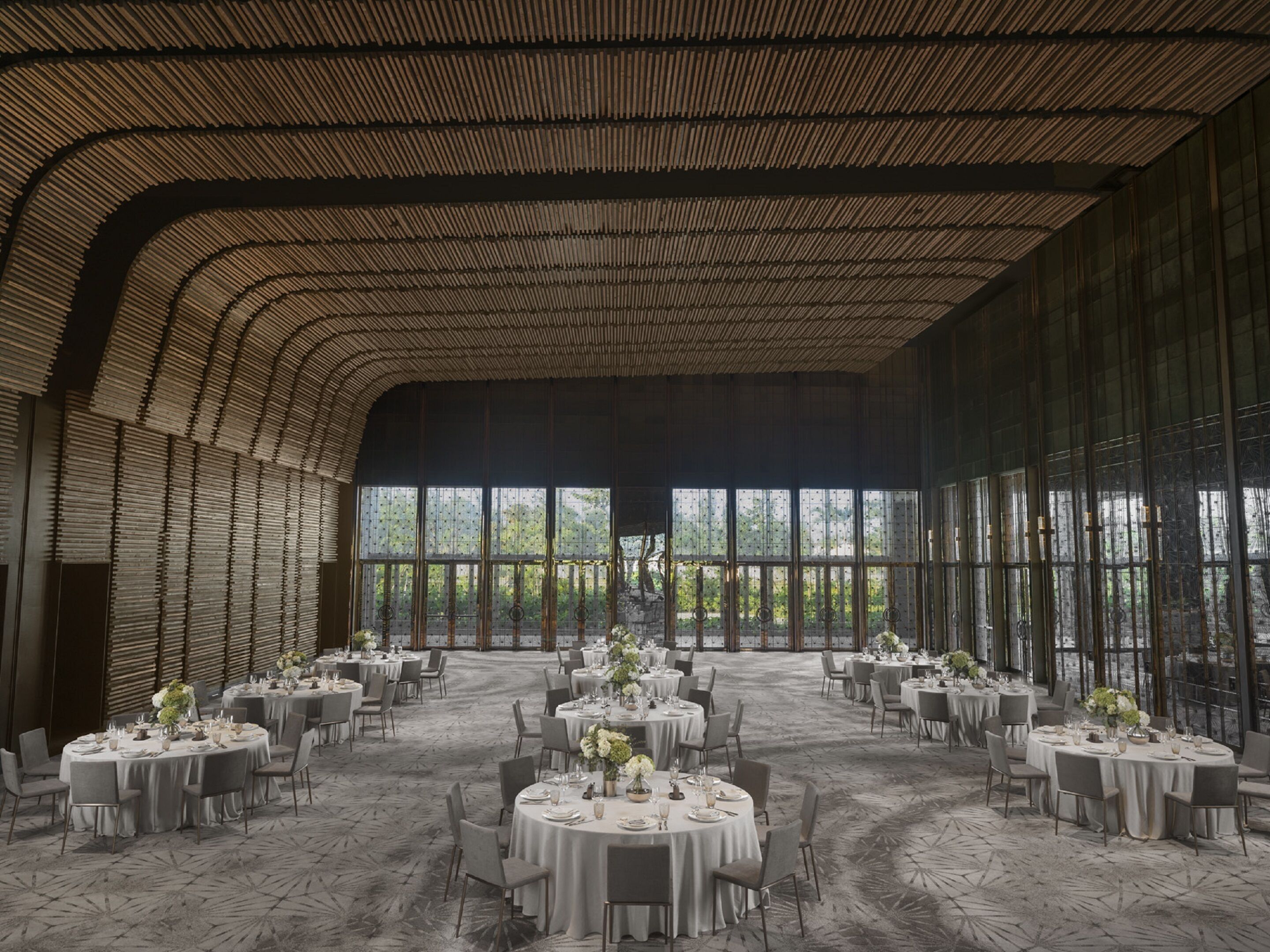 Un amplio salón de banquetes con mesas redondas dispuestas para comer, grandes ventanales que ofrecen vistas a zonas verdes y un techo alto con un diseño de madera único.
