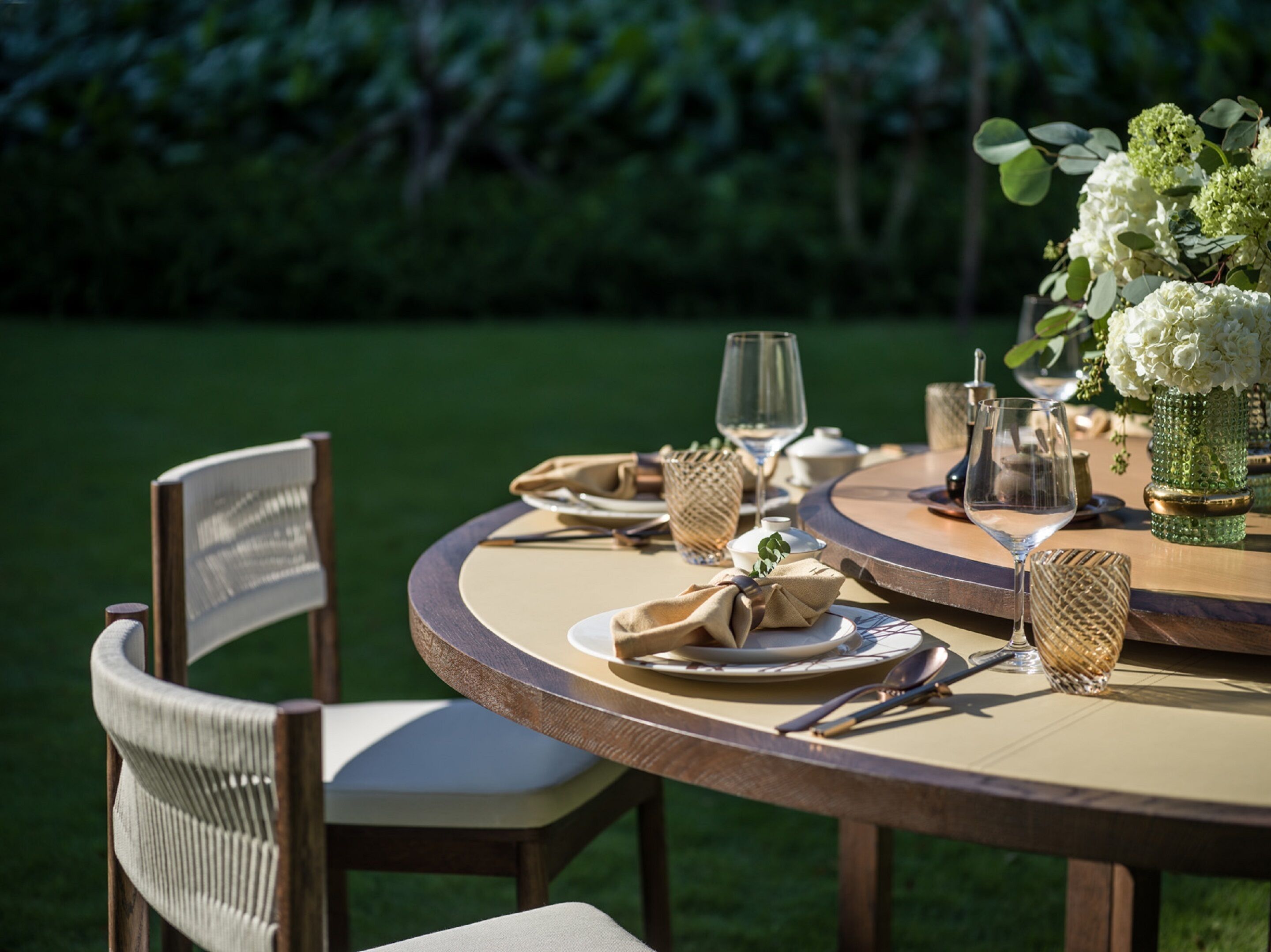 A round wooden table set for dining outdoors with chairs, plates, cutlery, and a vase of flowers.