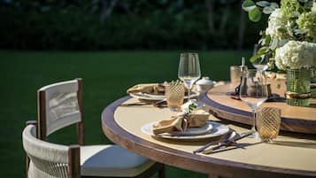 A round wooden table set for dining outdoors with chairs, plates, cutlery, and a vase of flowers.