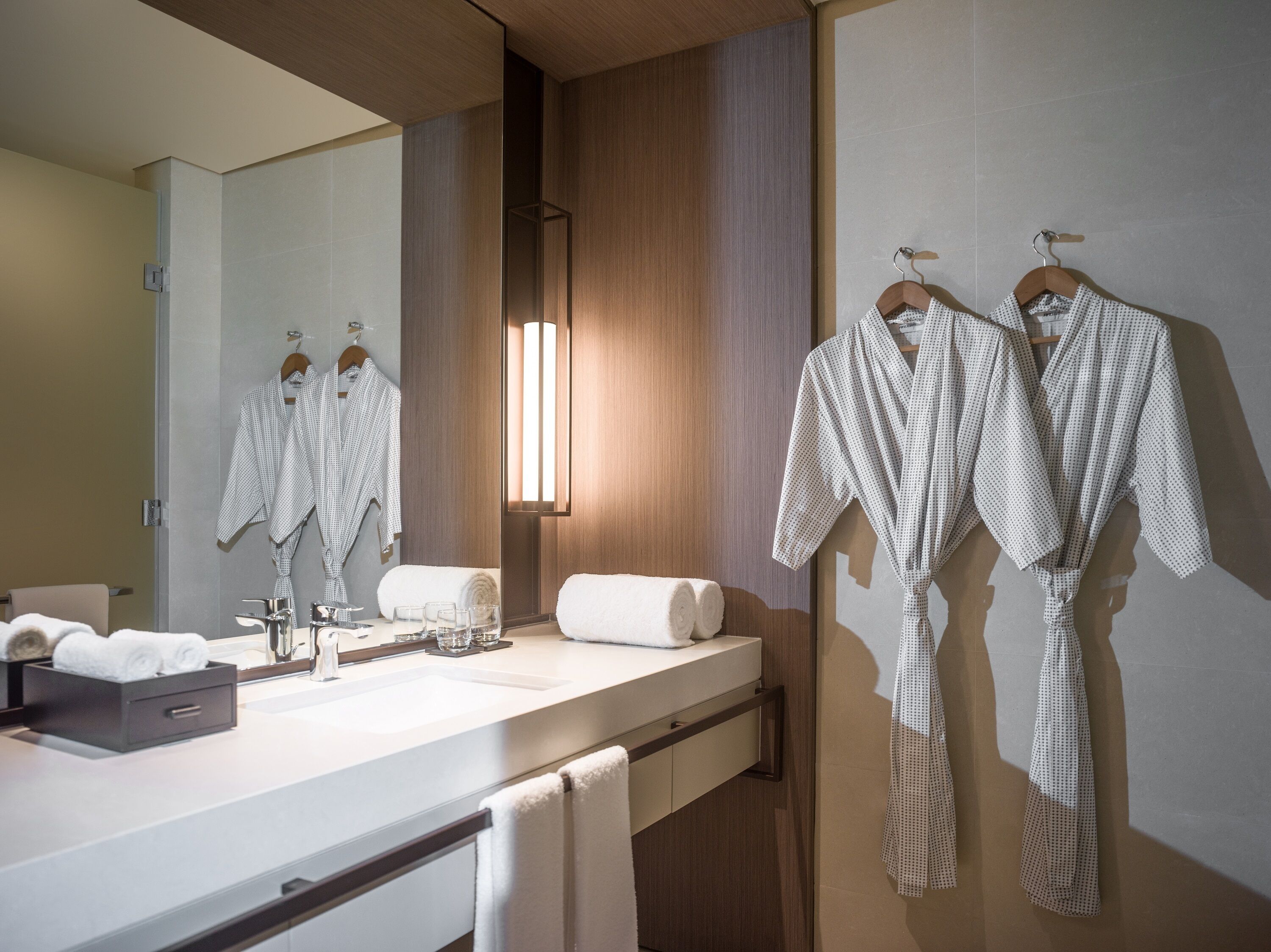 Un baño moderno con un espejo grande, paneles de madera, un lavamanos blanco y dos albornoces colgados en un perchero.