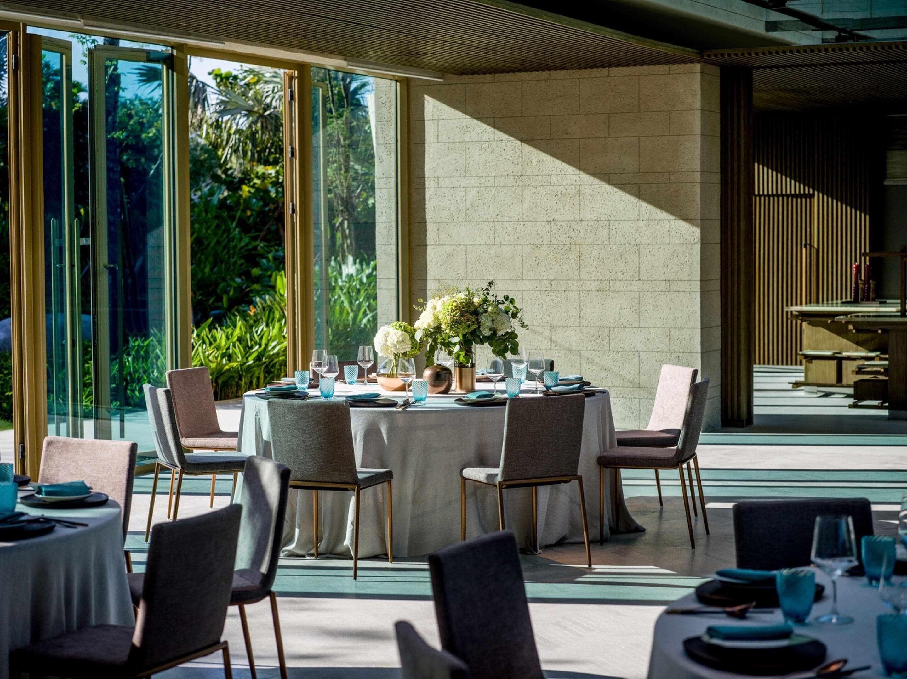 A dining area with a round table set for six, surrounded by chairs, with a view of greenery outside.