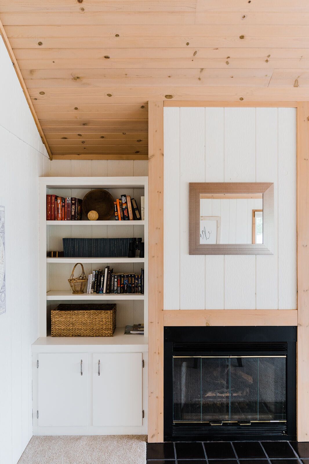 A room with a wooden ceiling, white walls, a fireplace, and built-in shelving with books and baskets. - Treloar Lakeside Cottage (Allegan)
