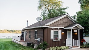 A small, single-story house with a porch, a satellite dish, and a view of a lake. - Treloar Lakeside Cottage (Allegan)