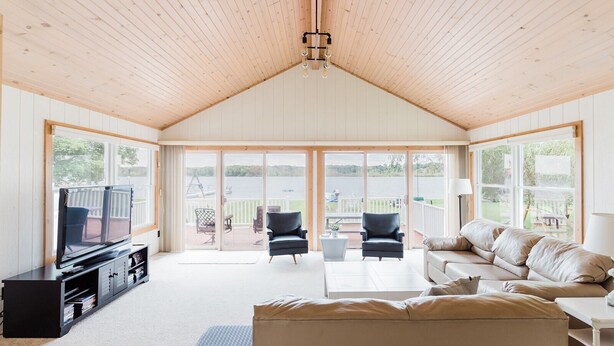 A spacious living room with a large TV, comfortable seating, and a view of a lake through large sliding glass doors. - Treloar Lakeside Cottage (Allegan)