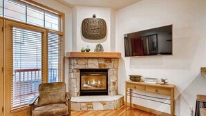 Un salon avec une cheminée en pierre, une horloge, un téléviseur fixé au mur et un fauteuil en bois.