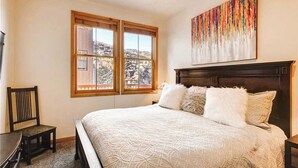 Une chambre à coucher avec un grand lit, deux fenêtres donnant sur un beau paysage, une tête de lit en bois et un tableau accroché au mur.