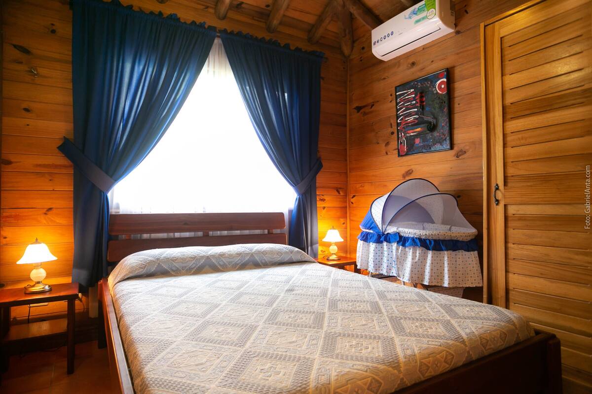A wooden cabin bedroom with a bed, two bedside lamps, a window with curtains, and a wall-mounted air conditioner.
