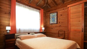 Un dormitorio de cabaña de madera con una cama, dos mesitas de noche, una mesita auxiliar, una silla y una puerta.