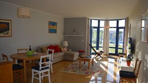 A well-lit living room with a dining area, a sofa, and a large window offering a view of the outdoors. - Luxury 2-bed apartment with huge pool - Orada resort (Albufeira)