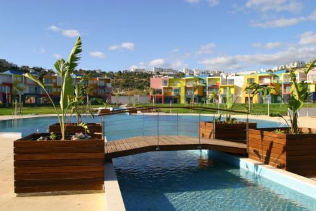 Una piscina con plataforma de madera y macetas, rodeada de edificios coloridos.